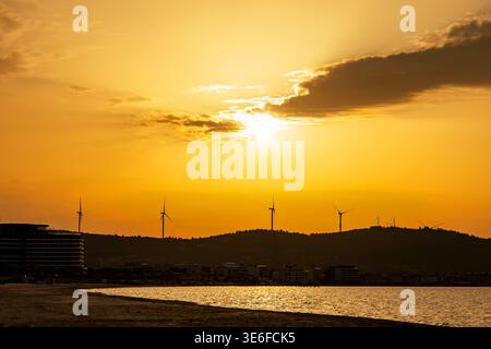 Der goldene Sonnenuntergang leuchtet über einem ruhigen Meer und fernen Hügeln der Küste, mit Windturbinen und Küstengebäuden in Silhouette unter dramatischen Wolken und W Stockfoto