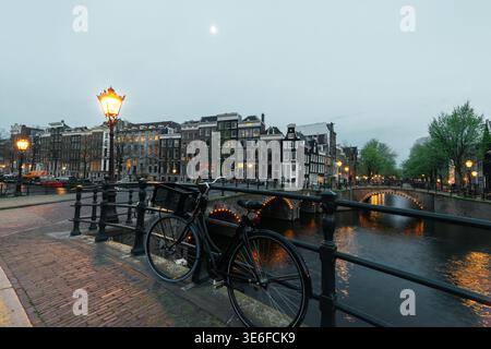Blick bei Dämmerung auf den Amsterdamer Kanal mit einem geparkten Fahrrad, leuchtenden Straßenlaternen, Bogenbrücken und historischen Kanalhäusern, die sich im ruhigen Wasser spiegeln, Niederlande Stockfoto