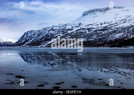 Ruhiger Winterfjord in der Nähe von Tromso, Norwegen, mit schneebedeckten Bergen Lapplands, die sich auf eisigem Wasser unter hellem Himmel spiegeln und eine ruhige arktische Landschaft schaffen Stockfoto
