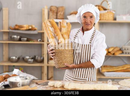 Glückliche, reife Bäckerin posiert mit Korb frisch gebackener Baguettes in der Bäckerei und demonstriert das Konzept der Lebensmittelindustrie und des Bäckerberufs Stockfoto