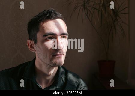 Porträt eines nachdenklichen jungen Mannes, der drinnen im dramatischen Sonnenlicht sitzt, mit sanften Schatten und einer Zimmerpflanze im Hintergrund. Warmes, stimmungsvolles Bild mit in Stockfoto