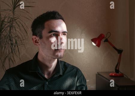 Innenporträt eines nachdenklichen jungen Mannes, der wegschaut, während er in einem sanft beleuchteten Raum mit einer roten Schreibtischlampe sitzt. Atmosphärisches Lifestyle-Bild mit Wärme Stockfoto