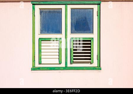 Traditionelles Holzfenster mit grünem Rahmen und weißen Fensterläden an einer pinkfarbenen Wand in Willemstad Curacao unter hellem Sonnenlicht Stockfoto