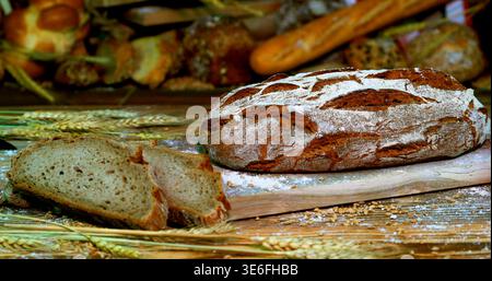 Frisch gebackenes Brot auf einem Holzbrett mit Weizen und verschiedenen Backwaren im Hintergrund bei Tageslicht Stockfoto