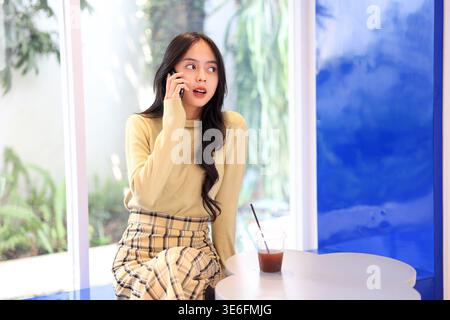 Asiatische junge Frau spricht oder telefoniert, während sie mit Americano auf einem Holztisch im Café sitzt. Freizeit Freizeit, Entspannung Im Freien, Urbanes Leben. Stockfoto