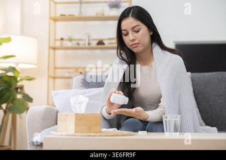 Eine junge Frau sitzt auf einer Couch in einem gemütlichen Wohnzimmer und nimmt Medikamente aus einer Flasche. Sie scheint sich unwohl zu fühlen, mit einer Decke, die um ihren Shou gewickelt ist Stockfoto