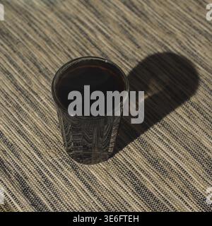 Eine Nahaufnahme fängt ein Glas mit sattem schwarzem Kaffee ein und wirft einen faszinierenden Schatten auf eine gewebte Textiloberfläche Stockfoto