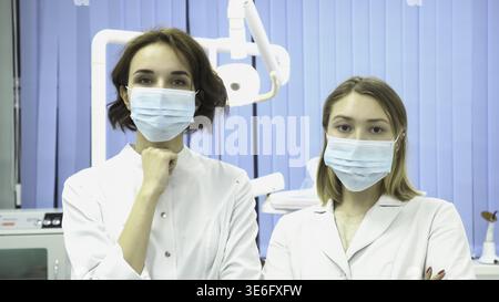 Zahnärztinnen mit Masken auf ihren Gesichtern blicken direkt auf die Kamera. Schöne junge Ärztinnen, die Uniform tragen und im Zahnschrank stehen, ich Stockfoto