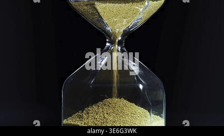 Goldener Sand in transparenter Sanduhr isoliert auf schwarzem Hintergrund. Kleine Metallpartikel, die durch eine Sanduhr laufen und die Verlaufszeit messen Stockfoto