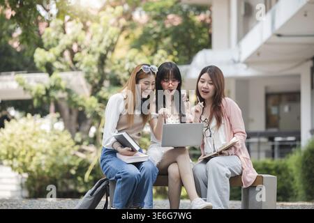 Eine Gruppe glücklicher junger asiatischer Studenten, die auf einer Bank sitzen, auf einen Laptop-Bildschirm schauen, über ihr Schulprojekt diskutieren und Brainstorming durchführen Stockfoto