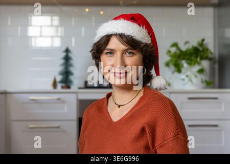 Frau posiert in der modernen Küche mit Weihnachtsmann-Hut und Roststrick-Pullover, zeigt Halskette Stockfoto