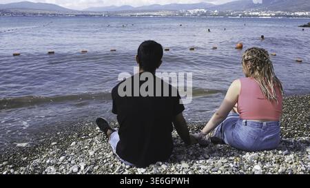 Das Paar hält die Hände, während es auf den Kieselsteinen in der Nähe des Meeres sitzt. Hinteres Porträt eines jungen Touristen-Paares, das sich mit zwei Händen am Kiesstrand hält Stockfoto