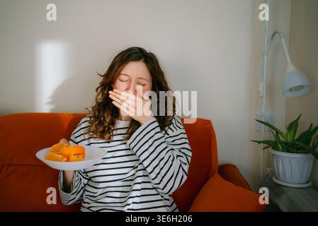 Junge Frau, die sich unwohl fühlt und unter starker Übelkeit leidet, den Mund bedeckt, während sie einen Teller Orangen hält, die Nahrungsabneigung und Toxikose erlebt Stockfoto