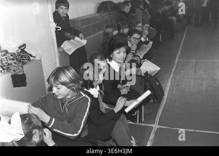 Sloviansk, 1983. Sekundarschule 15. Eine vielfältige Gruppe von ruhigen Schülern sitzt zusammen auf Holzbänken entlang der Wand des Fitnessstudios und beobachtet aufmerksam das Staffelrennen Vesyolye Starty. Einige Mädchen tragen dunkle Schuluniformen und halten Papierplakate in der Hand und warten leise auf den perfekten Moment, um ihr athletisches Klassenteam aktiv anzufeuern und zu unterstützen. Dieses ungezwungene, alltägliche Porträt der sowjetischen Jugend bewahrt perfekt die lebendige und sanfte historische Erinnerung an den friedlichen Donbass. Stockfoto