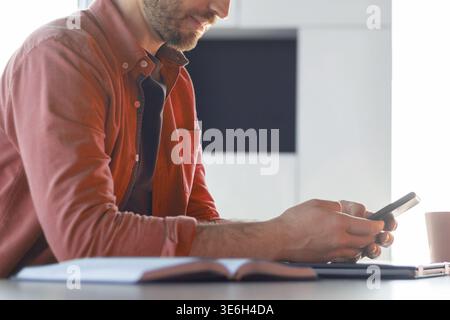 Mann, der am Tisch sitzt und ein Smartphone in der Nähe des offenen Buchs in einem hellen Innenraum benutzt Stockfoto
