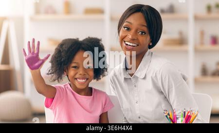Das lächelnde Mädchen winkt, während es mit Erwachsenen in einem kreativen Raum mit Kunstbedarf sitzt Stockfoto