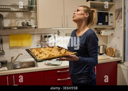 Frau mittleren Alters hält stolz ein Tablett mit frisch gebackenem goldenem Schinken und Käsegebäck in der Küche Stockfoto