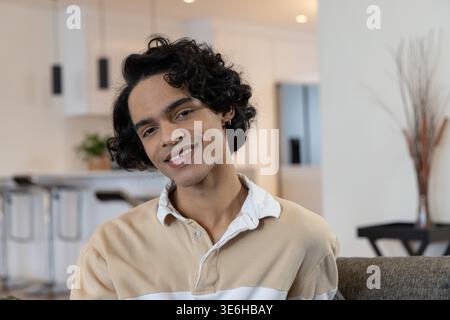 Mann Mitte 20 sitzend und lächelnd auf Sofa im offenen Wohnzimmer mit beigem Pullover Stockfoto