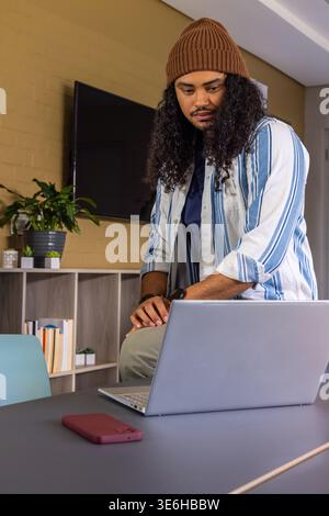Mann in brauner Beanie, der sich auf dem Tisch mit silbernem Laptop im Wohnzimmer mit rotem Smartphone lehnt Stockfoto