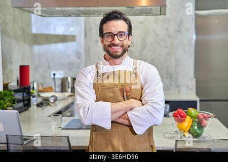 Selbstbewusster Koch in einer Küchenjacke und Schürze mit überkreuzten Armen, stehend und lächelnd in einer modernen Küche Stockfoto