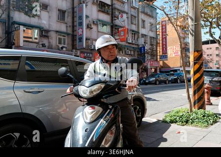 Taipei, Taiwan - 3. Februar 2024: Älterer Mann mit Helm fährt auf einem Bürgersteig mit dem Roller gegen den Verkehr auf einer sonnigen Stadtstraße Taipeis. Stockfoto