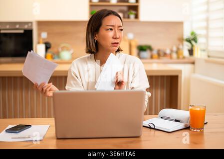 Eine Frau hält zwei Papiere und vergleicht sie mit Informationen auf ihrem Laptop, was Verwirrung und Besorgnis über Ausgaben oder Zahlungen zeigt. Stockfoto