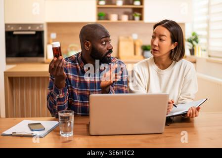 Ein besorgter Mann hält eine Pille-Flasche, während sein Partner in ein Notizbuch schreibt, was auf eine gesundheitliche Sorge zu Hause hindeutet. Stockfoto