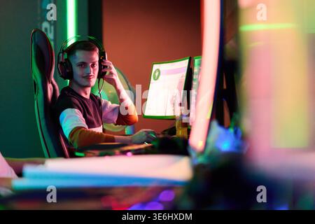 Ein fokussierter Spieler sitzt in einem Spielesessel mit Headset, streamt und spielt auf zwei Monitoren in einem neonbeleuchteten Spielzimmer und vermittelt Konzentration, Spannung und moderne E-Sport-Kultur. Stockfoto