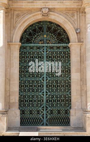 Grünes schmiedeeisernes Tor der San Bartolomeo Kirche in Scicli, eingerahmt von Steinsäulen und Bogen, mit floralen und geometrischen Mustern. Stockfoto