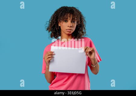 Ernste Frau, die ein weißes a4-Blatt Papier auf blauem Studiohintergrund hält. Protest, Beschwerde, Ankündigung, Nachricht, Frustration, Werbemockup, Kopierraum für Text oder Design. Hohe Qualität Stockfoto