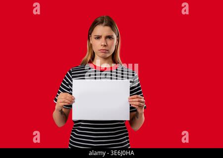 Ernste Frau, die ein weißes a4-Blatt Papier auf rotem Atelierhintergrund hält. Protest, Beschwerde, Ankündigung, Nachricht, Frustration, Werbemockup, Kopierraum für Text oder Design. Hohe Qualität Stockfoto