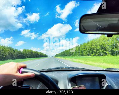 Ein Fahrer hält das Lenkrad auf einer Autobahn, umgeben von grünen Bäumen und einem hellblauen Himmel während der Fahrt. Stockfoto