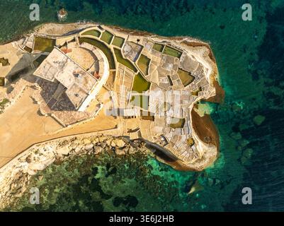 Aus der Vogelperspektive der geometrischen Salinen im Kontrast zum türkisfarbenen Meer, neben einem Gebäude auf einem Felsvorsprung, Iż-Żebbuġ, Gozo, Malta. Stockfoto