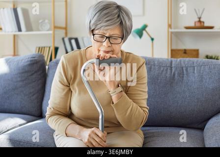 Ältere Frau mit Stock sitzt allein auf dem Sofa zu Hause, traurige Person denkt Stockfoto