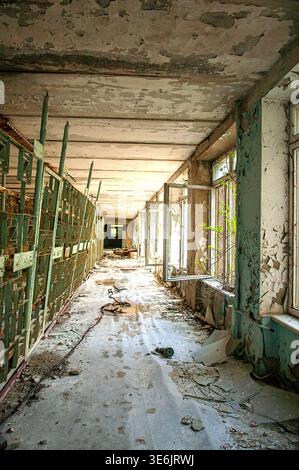Ukraine, Pripyat: Langer, verfallener Flur innerhalb einer verlassenen Schule mit abblätternder Decke und Wänden, grünen Metallgeländern, offenen Fenstern mit Vegetati Stockfoto