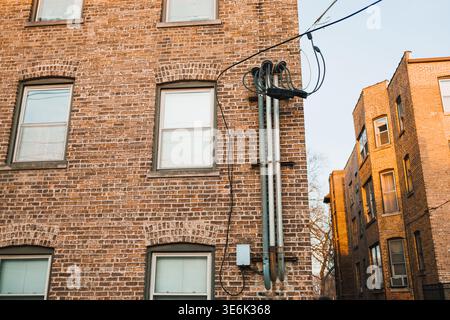 Kabel verlaufen entlang der Seite eines Ziegelgebäudes in der Nähe von zwei anderen Gebäuden. Die Sonne untergeht und wirft sanftes Licht auf die Ziegelwände und Fenster. Stockfoto