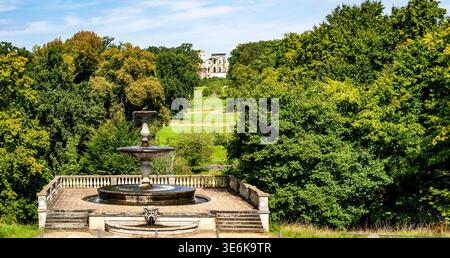 Kunstvoller Steinbrunnen auf der Terrasse mit Blick auf den üppigen grünen Park und das ferne Herrenhaus unter blauem Himmel. Stockfoto