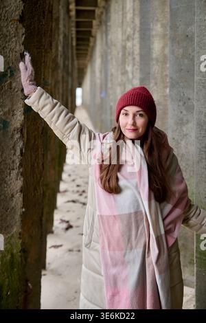 Erwachsene Frau mit langen braunen Haaren in beigefarbenem, gepolstertem Mantel, roter Strickmütze, rosa Handschuhen und großem kariertem rosa Schal, der die strukturierte Steinwand berührt Stockfoto