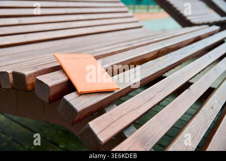 Orangefarbener Pass auf brauner Holzlattenbank im Park mit verschwommenem Hintergrund. Weiches natürliches Tageslicht beleuchtet strukturierten ben Stockfoto