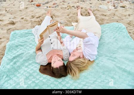 Zwei junge Frauen liegen auf einer helltürkisfarbenen Decke am Sandy Beach und lesen gemeinsam Buch bei weichem Tageslicht mit verstreuten Muscheln Stockfoto