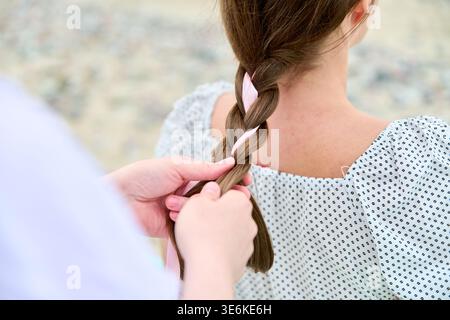 Erwachsene Frau mit braunem Haar in einer Bluse mit Punktmuster hat ihr Haar mit rosa Schleife von einer anderen Person im Freien geflochten. Das Bild zeigt die Hände, die Haar weben Stockfoto