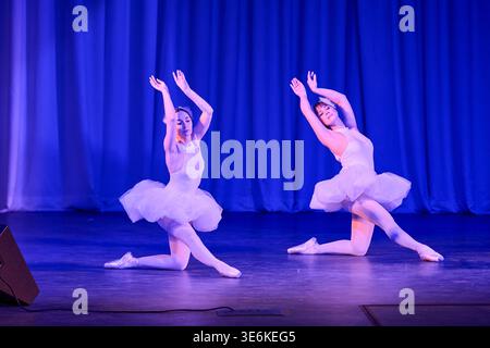 Anmutige Ballerinas in weißem Tutus, die kniende Posen mit erhobenen Armen auf der schwach beleuchteten Bühne vor tiefblauen Vorhängen ausführen und Eleganz und Eleganz in Szene setzen Stockfoto