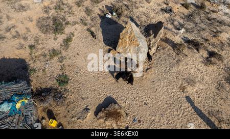 Von oben aus blickt man auf einen Wüstencamping mit einem provisorischen Leinwandzelt neben einem großen Felsbrocken, Ziegenessen, Fußabdrücke im Sand und verstreuten Gegenständen A Stockfoto