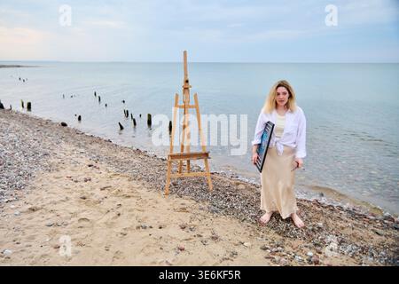 Junge Erwachsene Frau mit blonden Haaren läuft barfuß auf felsigem Sandstrand am ruhigen Meer, die gerollte Leinwand in der Nähe der Holzstaffelei unter weichem Tageslicht und Clos hält Stockfoto