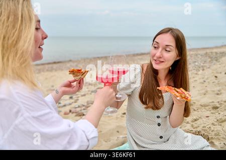 Zwei junge Frauen teilen sich Pizza und Toast mit rosafarbenem Wein, während sie am Sandstrand unter bewölktem Himmel sitzen und entspannte und fröhliche Momente zusammen genießen Stockfoto