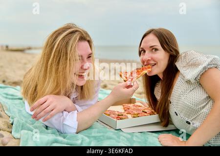 Zwei junge Frauen teilen sich ein Stück Pizza, während sie tagsüber auf der Decke am Strand entspannen und fröhliche und ungezwungene Momente auf dem Meer festhalten Stockfoto
