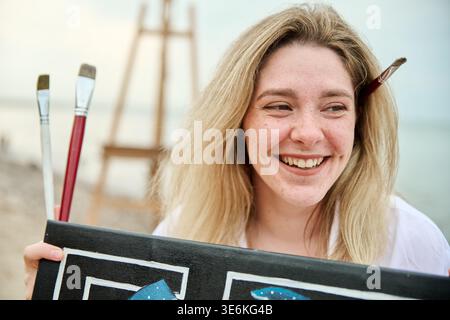 Die junge Erwachsene Malerin mit blonden Haaren und Sommersprossen lächelt fröhlich, während sie die Leinwand hält. Die Staffelei des Kunststudios ist im Hintergrund verschwommen Stockfoto