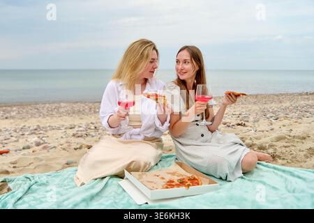 Zwei junge Frauen sitzen auf einer blauen Decke am Strand, teilen sich Pizza und rosa Wein unter bewölktem Himmel und genießen entspannte und fröhliche Momente zusammen Stockfoto