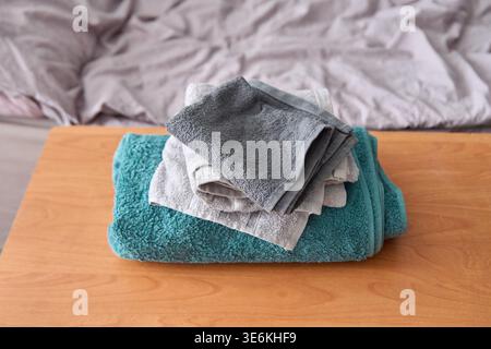 Weiche, strukturierte graue und beige Handtücher, die ordentlich gefaltet und auf dickem blaublauem Handtuch gestapelt sind, das auf einem Holztisch neben dem Bett mit hellrosafarbener Bettwäsche gestellt wurde Stockfoto