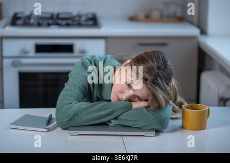 Erschöpfte junge Frau, die müde ist, Frustration während der Arbeit oder beim Lernen von zu Hause aus, mit dem Kopf auf einem Laptop am Küchentisch liegend und mit zehn schaut Stockfoto